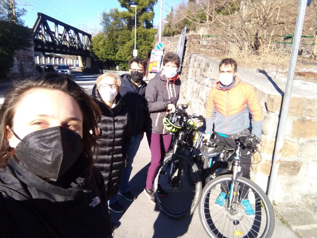 Selfie di gruppo in bici con mascherine vicino a un ponte soleggiato.