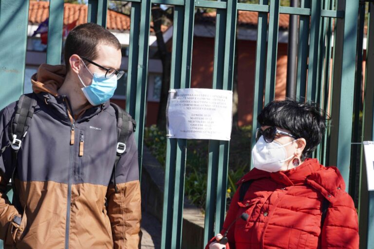 Due persone mascherate davanti a un cancello verde con avviso, in strada.