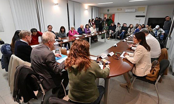 Riunione affollata in sala conferenze professionale con persone attorno a tavolo ovale.