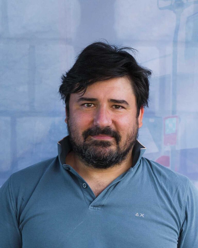 Ritratto professionale di uomo con barba, polo blu e sfondo azzurro sfumato.