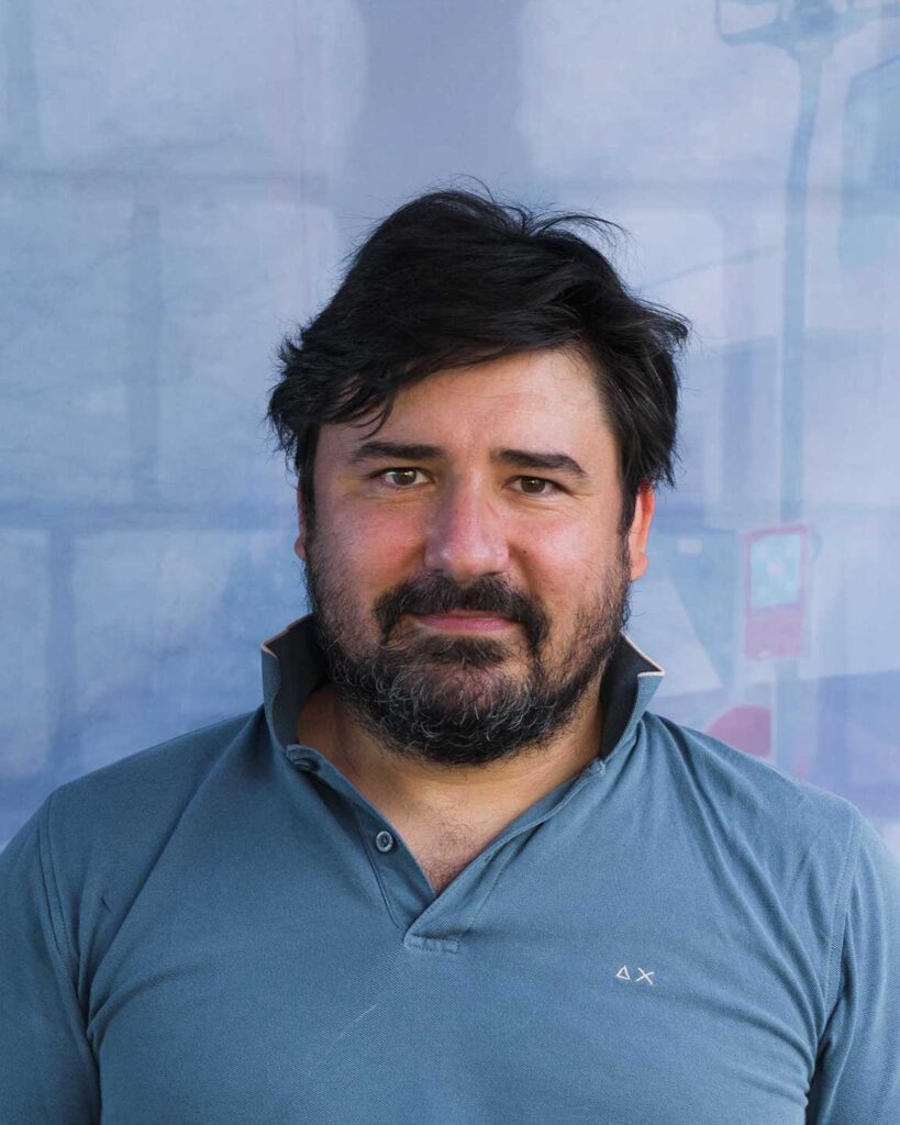 Ritratto professionale di uomo con barba, polo blu e sfondo azzurro sfumato.