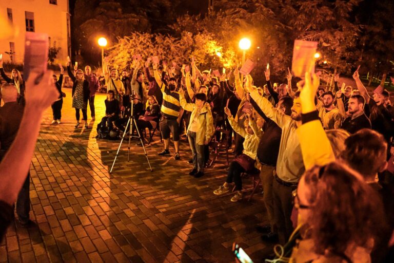 Protesta notturna in piazza con folla a braccia alzate sotto luci calde.