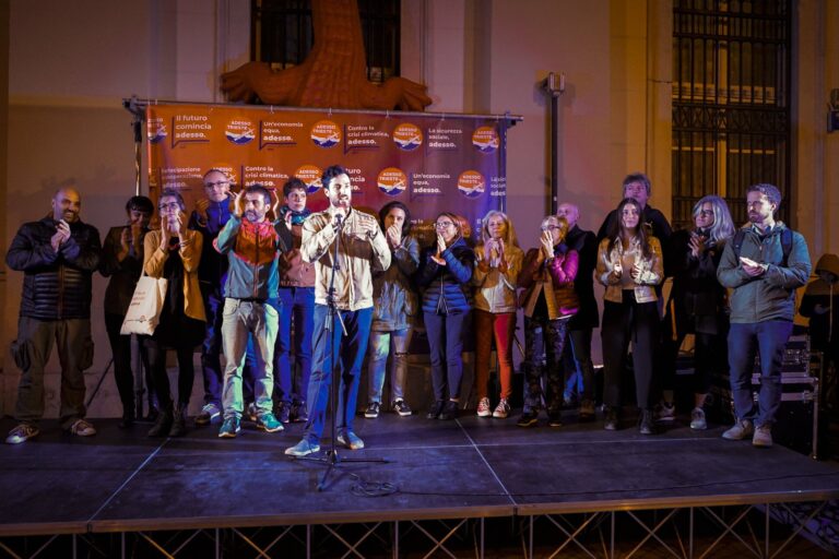 Evento notturno su palco: discorso al microfono, applausi e fondale arancione.