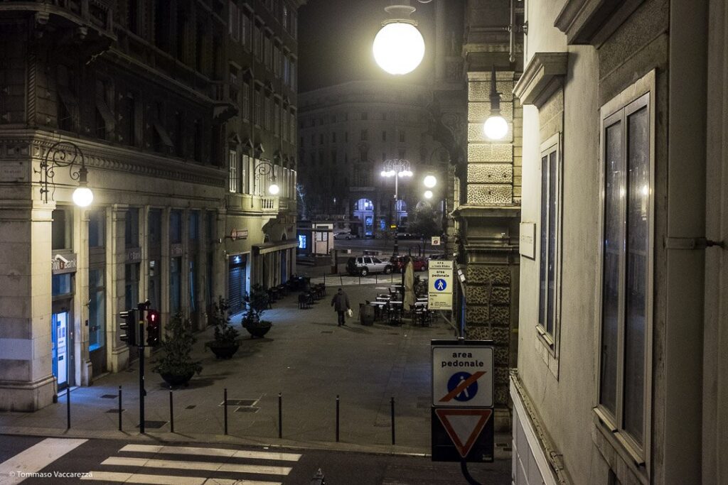 Strada notturna europea con lampioni, palazzi storici e pochi pedoni.