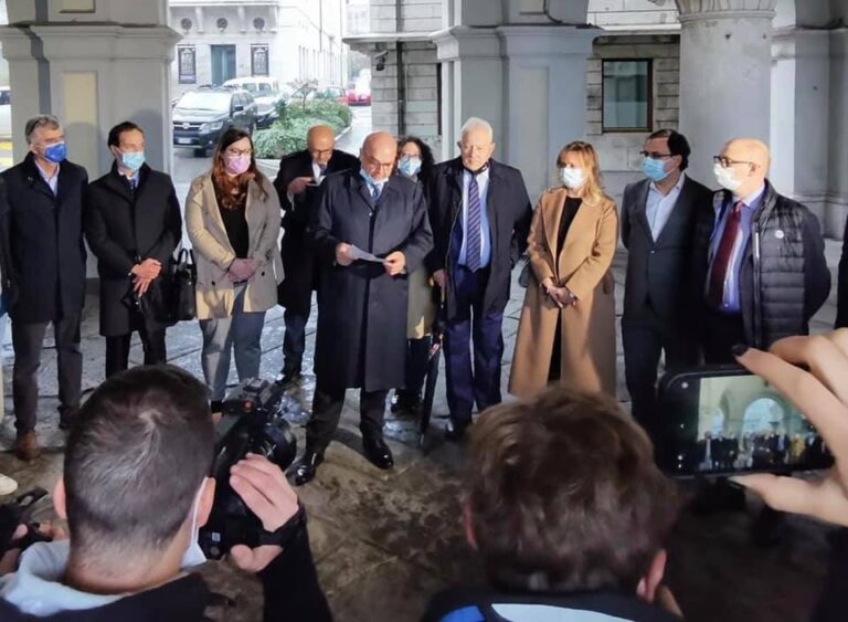 Conferenza stampa notturna davanti a palazzo storico con ufficiali mascherati e giornalisti.
