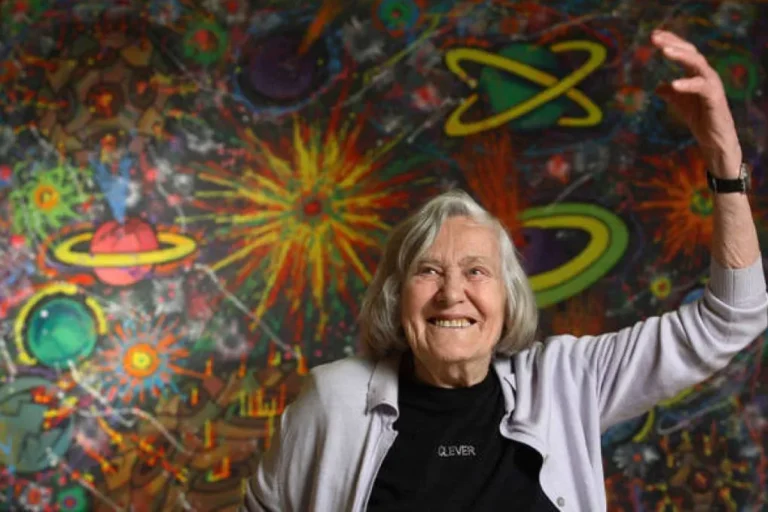 Margherita Hack sorride davanti a un murale cosmico colorato, con le braccia alzate.