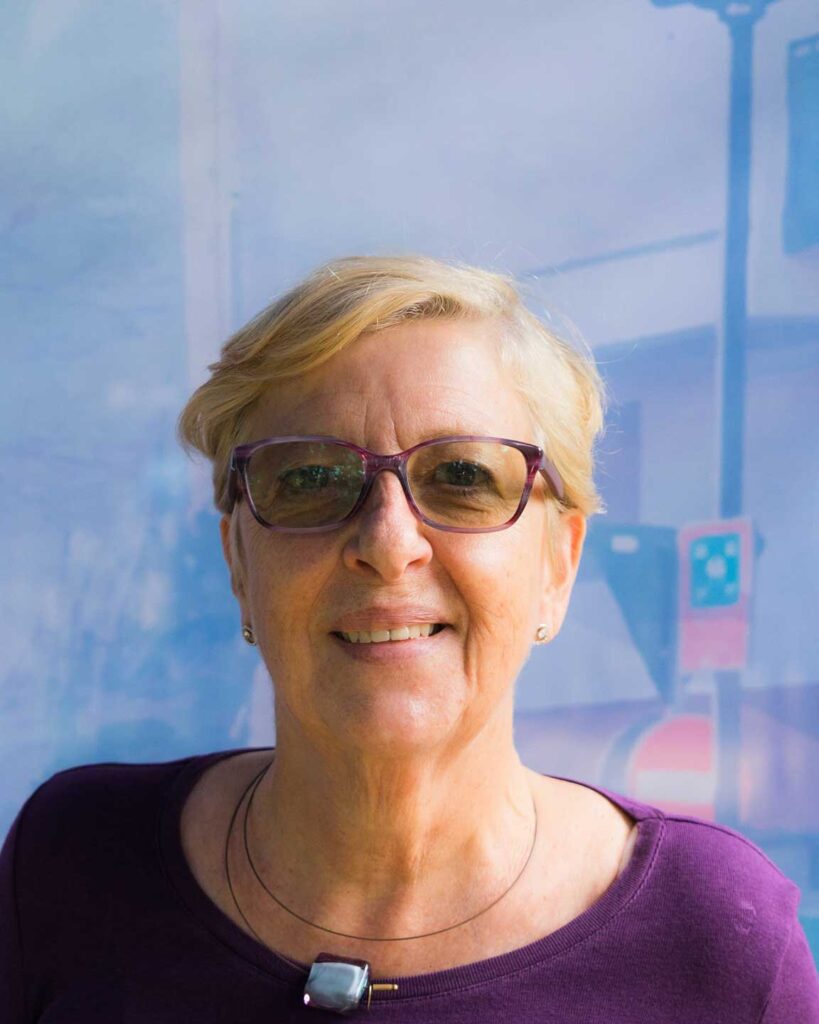 Ritratto di donna anziana con occhiali, sorriso lieve e sfondo sfocato.