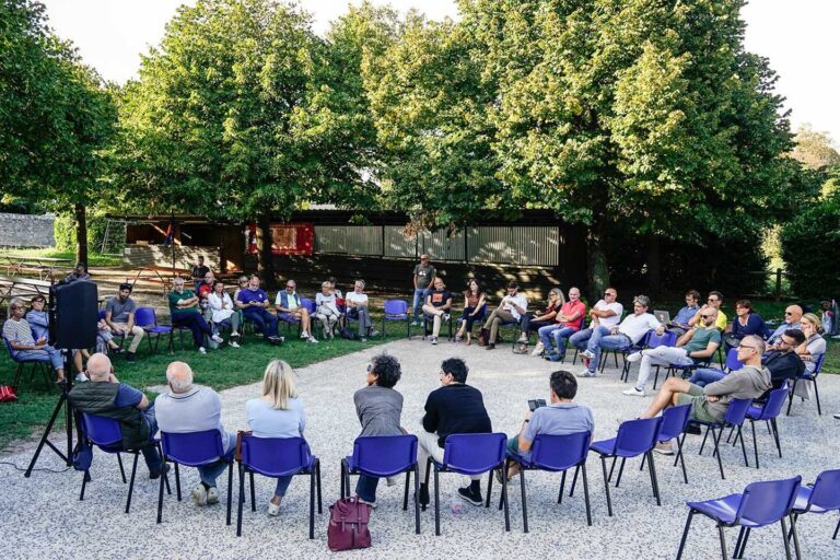 Incontro comunitario all’aperto in giardino, persone sedute in cerchio per discussione.