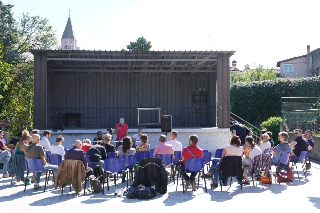 Pubblico all’aperto assiste a spettacolo comunitario su palco in legno, tra alberi e sole.