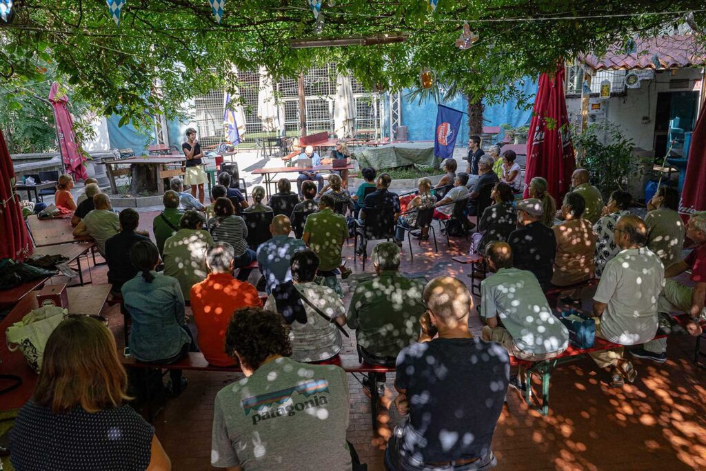Incontro conviviale all’aperto in cortile ombreggiato con tavoli, ombrelloni rossi e pubblico riunito.