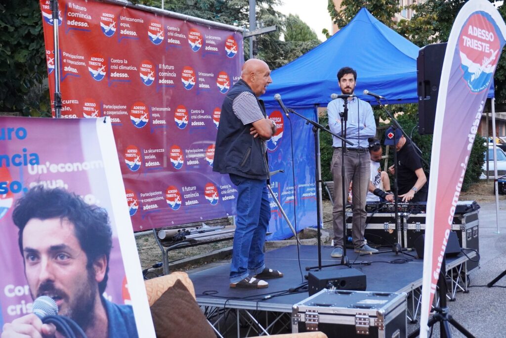 Uomo parla al microfono su palco all’aperto con backdrop brandizzato e gazebo blu.