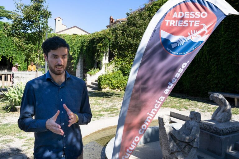 Uomo parla in giardino con banner Adesso Trieste durante presentazione all’aperto.
