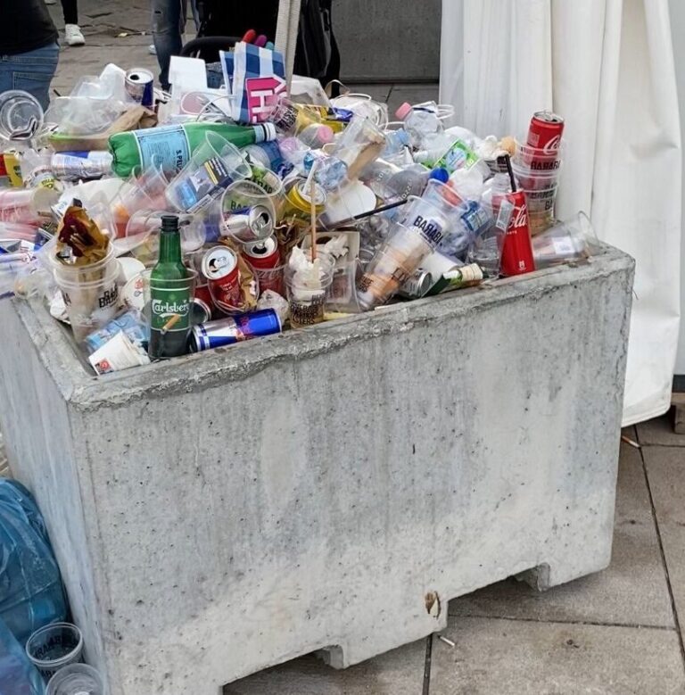Bidone dei rifiuti in cemento strabordante all’aperto, pieno di bottiglie, bicchieri e imballaggi.