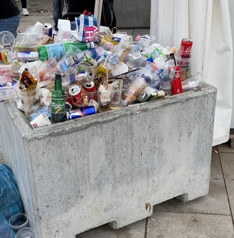 Bidone dei rifiuti in cemento strabordante all’aperto, pieno di bottiglie, bicchieri e imballaggi.