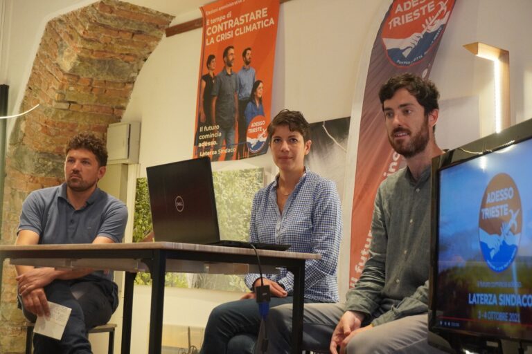 Panel di discussione con tre relatori, laptop, poster e monitor in sala conferenze.