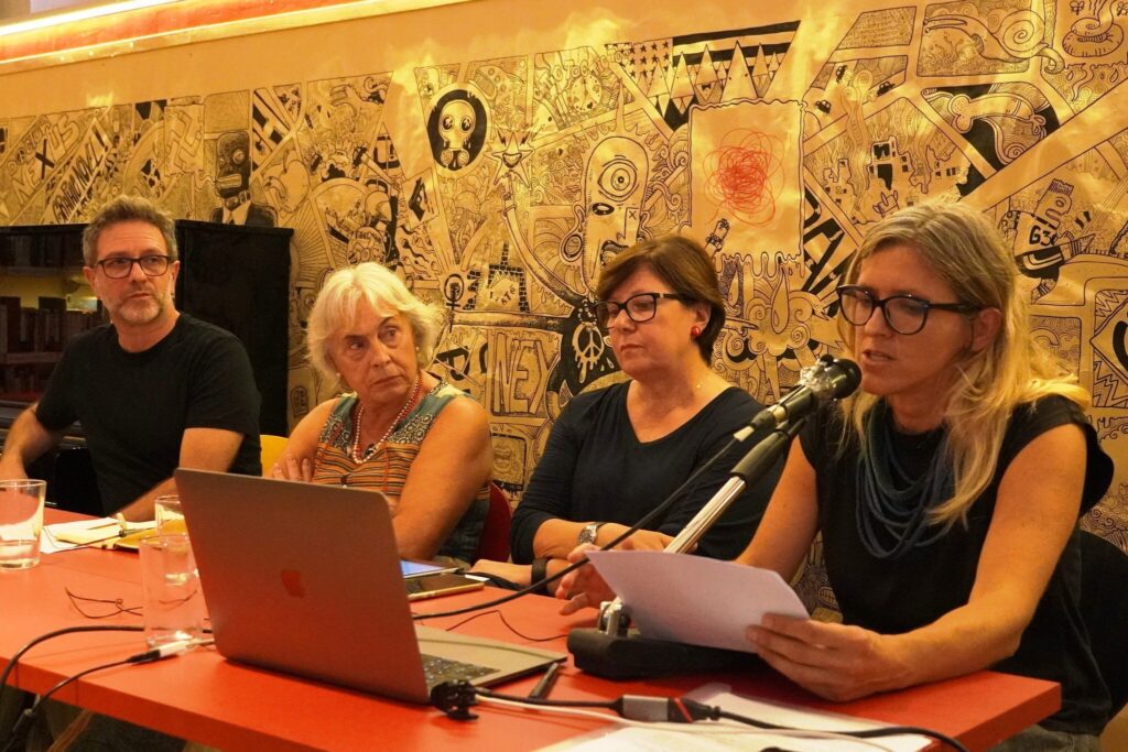 Panel a Trieste con quattro relatori, microfoni e murale artistico colorato sullo sfondo.