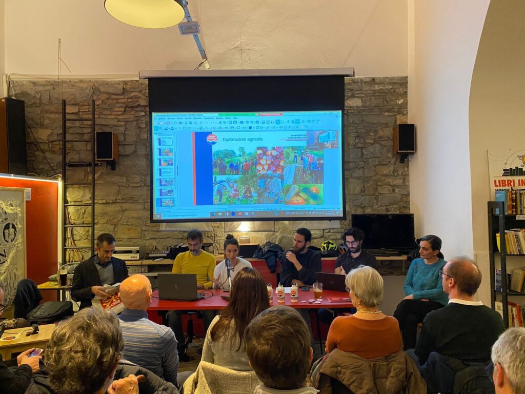 Tavola rotonda in libreria con pubblico, relatori al tavolo e slide proiettata.