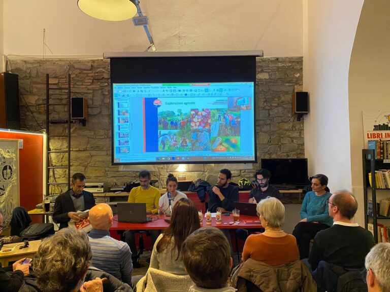 Tavola rotonda in libreria con pubblico, relatori al tavolo e slide proiettata.