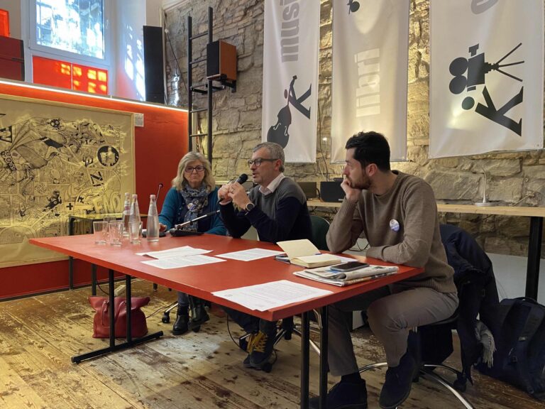Tavola rotonda medievale con tre relatori, microfono, tavoli rossi e muri in pietra.
