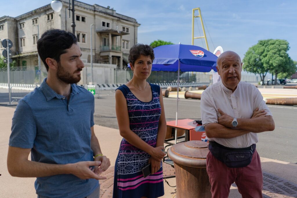 Tre persone in piazza vicino a un chiosco con ombrellone blu.