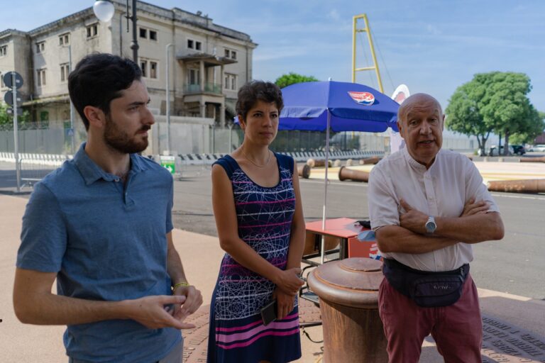 Tre persone in piazza vicino a un chiosco con ombrellone blu.