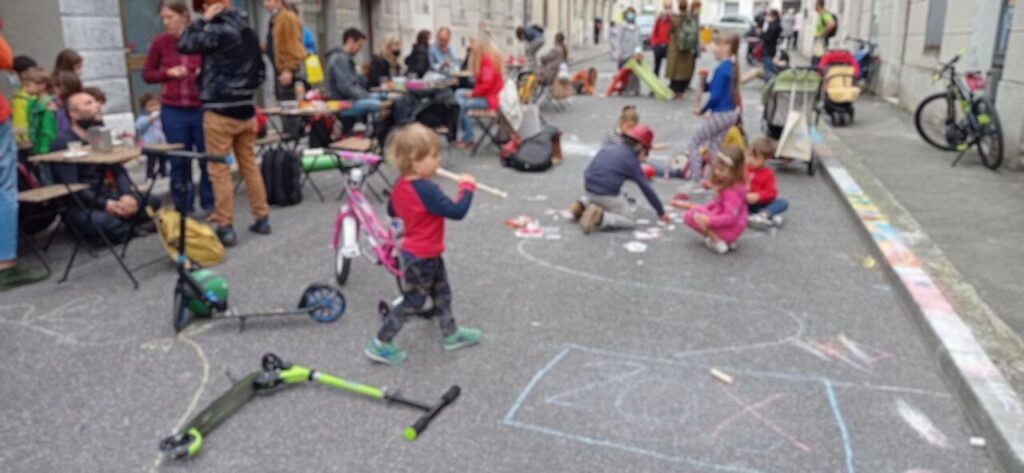 Bambini giocano con gessetti e monopattini in una strada chiusa al traffico.