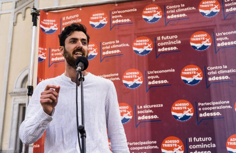 Uomo parla al microfono a Trieste davanti al backdrop politico Adesso.