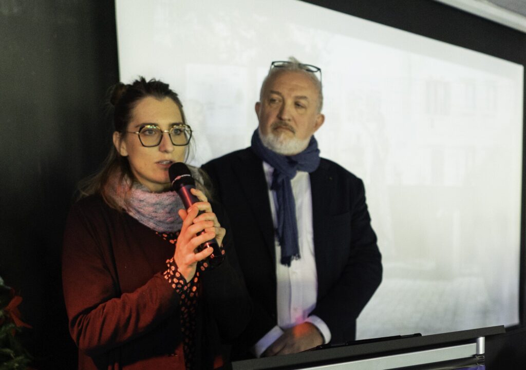 Relatrice al microfono con collega durante conferenza davanti a schermo di proiezione.