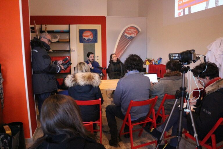 Conferenza stampa con pubblico, relatore al tavolo e telecamera su treppiede.
