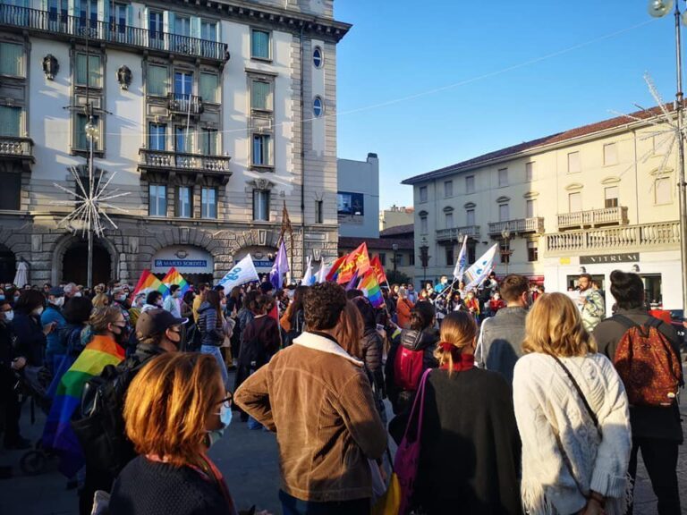 Pride in piazza storica europea con folla e bandiere arcobaleno sotto cielo azzurro.