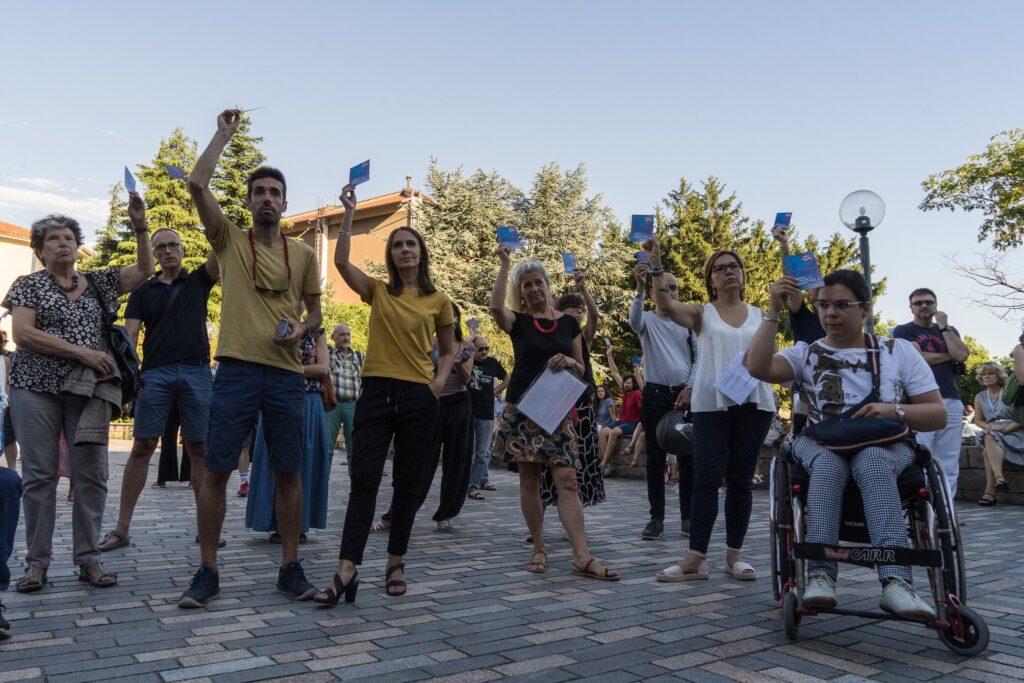 Corteo solidale all’aperto con bandiere gialloblu e persona in sedia a rotelle.