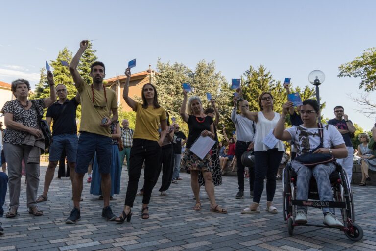 Corteo solidale all’aperto con bandiere gialloblu e persona in sedia a rotelle.