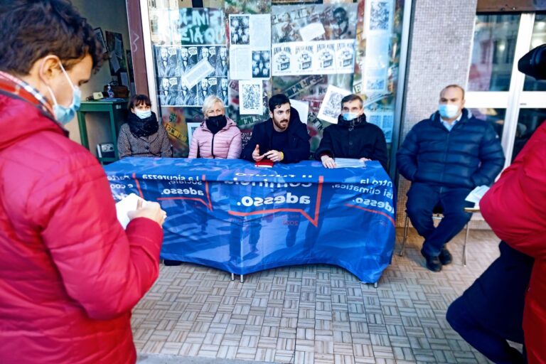 Protesta con banner blu e mascherine davanti a un edificio pubblico.