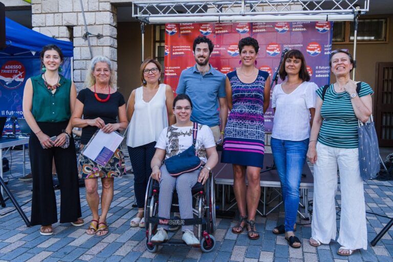 Gruppo sorridente a evento pubblico all’aperto con donna in sedia a rotelle.