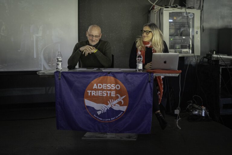 Incontro pubblico Adesso Trieste: due relatori, banner blu, microfono e laptop.