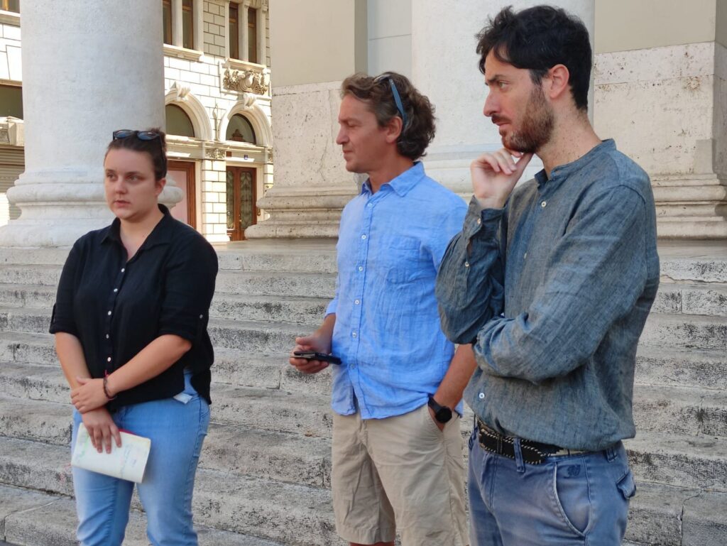 Tre persone discutono sui gradini di un palazzo storico pubblico.