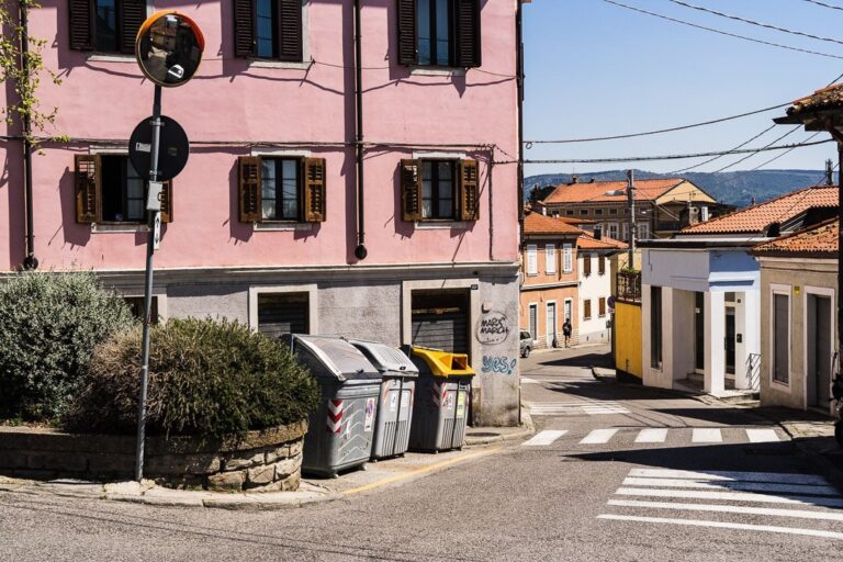 Angolo di borgo soleggiato con casa rosa, persiane verdi e colline sullo sfondo.
