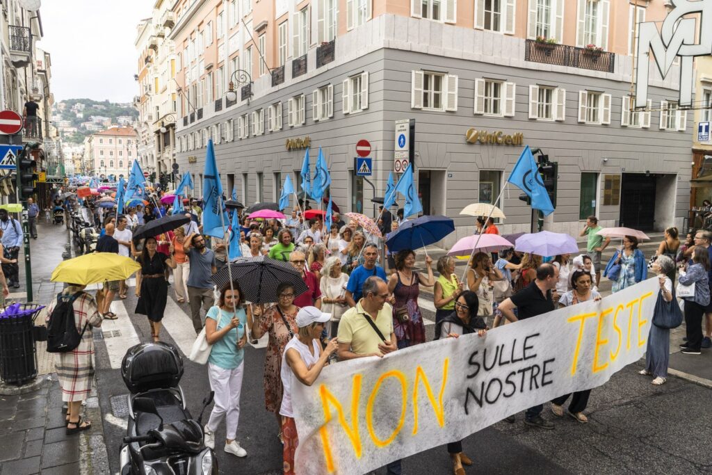 Manifestazione sotto la pioggia con bandiere blu e striscione “NON SULLE NOSTRE...” in città.