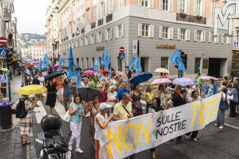 Manifestazione sotto la pioggia con bandiere blu e striscione “NON SULLE NOSTRE...” in città.