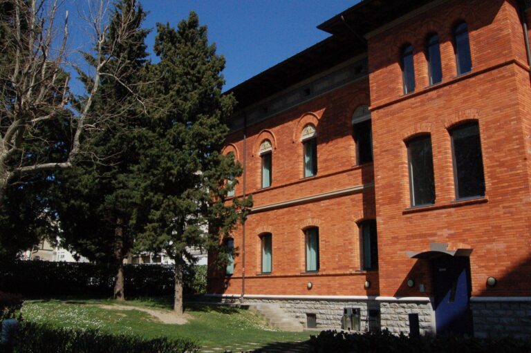 Edificio storico in mattoni rossi nel campus, con finestre ad arco e alberi.