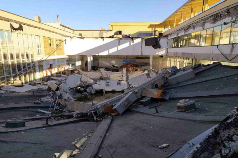 Crollo del tetto tra edifici industriali con detriti, travi piegate e gravi danni.
