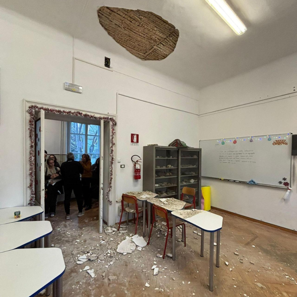 Crollo del soffitto in aula scolastica con detriti, banchi spostati e persone all’ingresso.