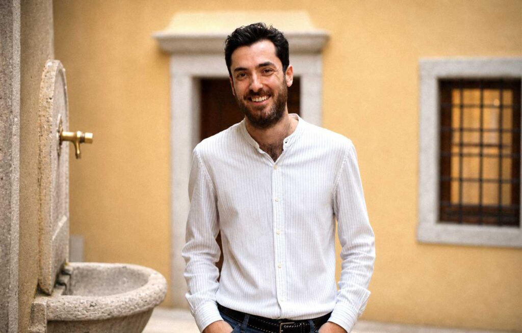 Ritratto di uomo sorridente in cortile mediterraneo, camicia bianca e fontana in pietra.