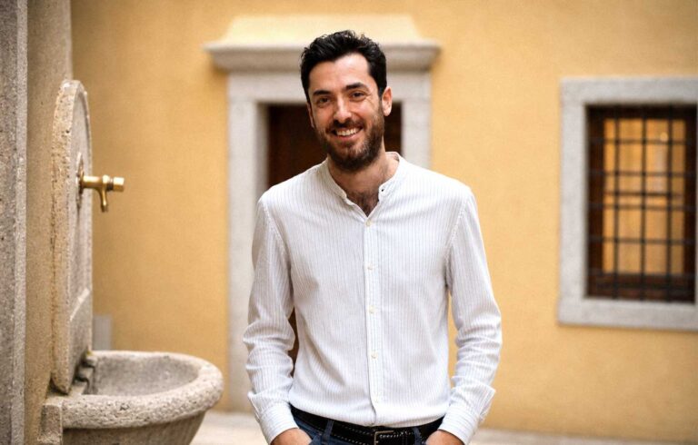 Ritratto di uomo sorridente in cortile mediterraneo, camicia bianca e fontana in pietra.