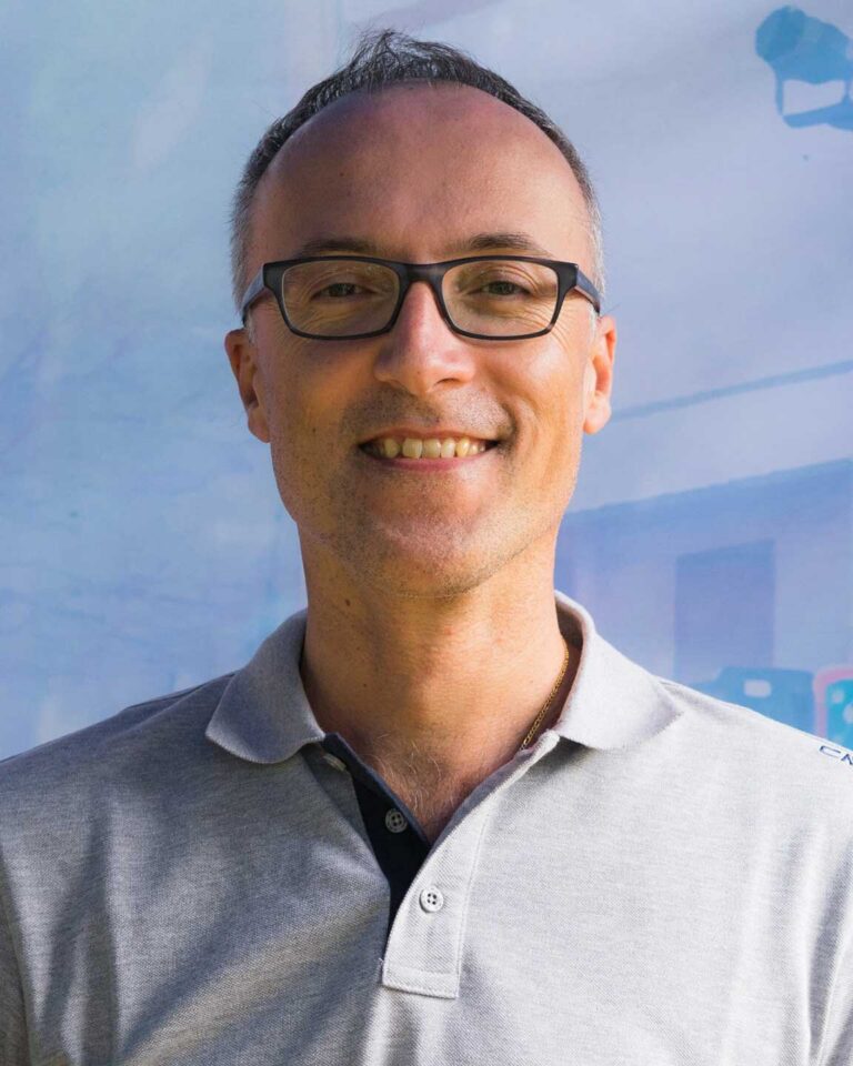 Ritratto professionale di uomo sorridente con occhiali neri e polo grigia.