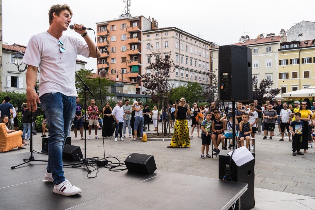 Artista di strada canta su palco all’aperto in piazza urbana davanti a un piccolo pubblico.
