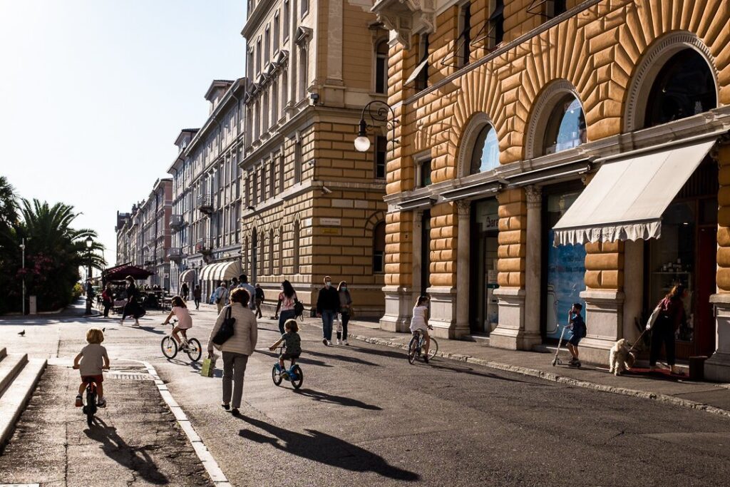 Strada europea assolata con bambini in bici, palazzi storici e ombre lunghe.
