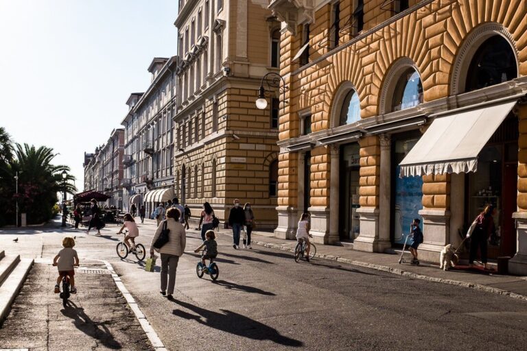 Strada europea assolata con bambini in bici, palazzi storici e ombre lunghe.