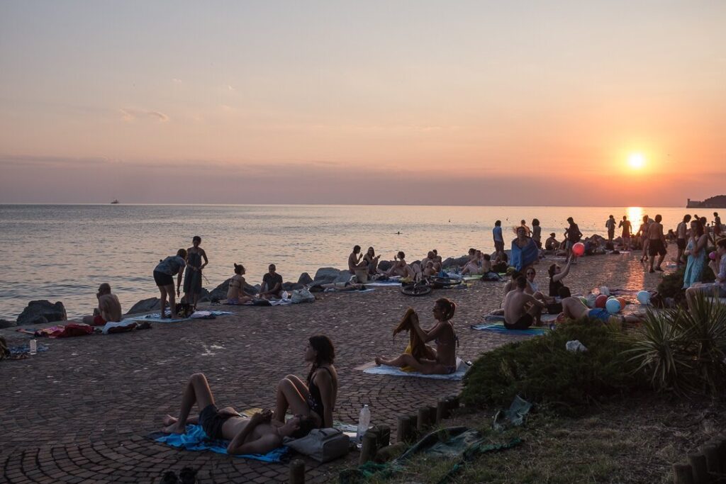 Spiaggia al tramonto con mare calmo, folla sulla riva e sole basso all’orizzonte.