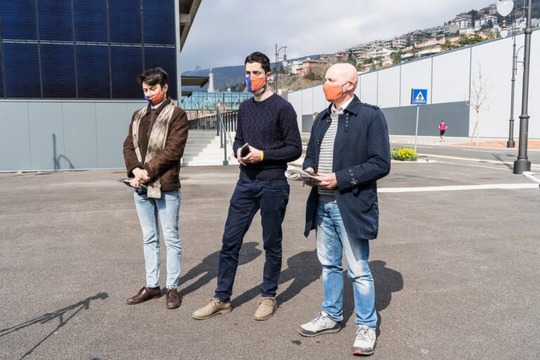 Tre uomini in briefing all’aperto davanti a edificio moderno e piazzale asfaltato.